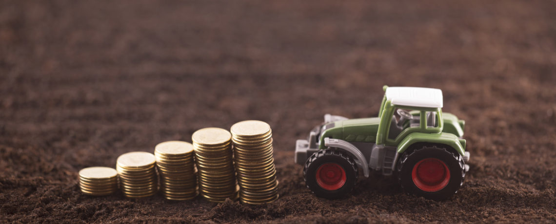 A miniature tractor and an ascending stack of coins rest on a plowed field.