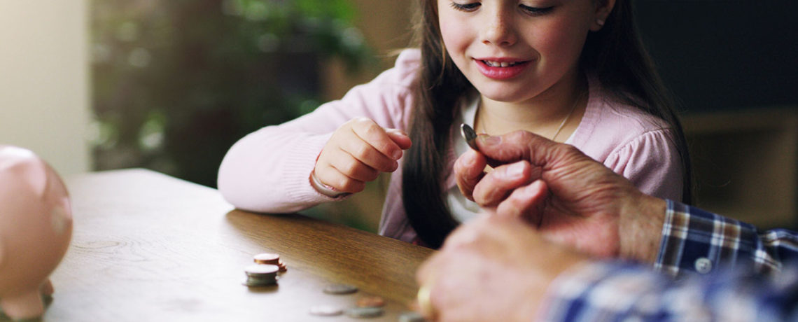 An older man teaches a young girl about financial literacy at a table with coins and a piggy bank