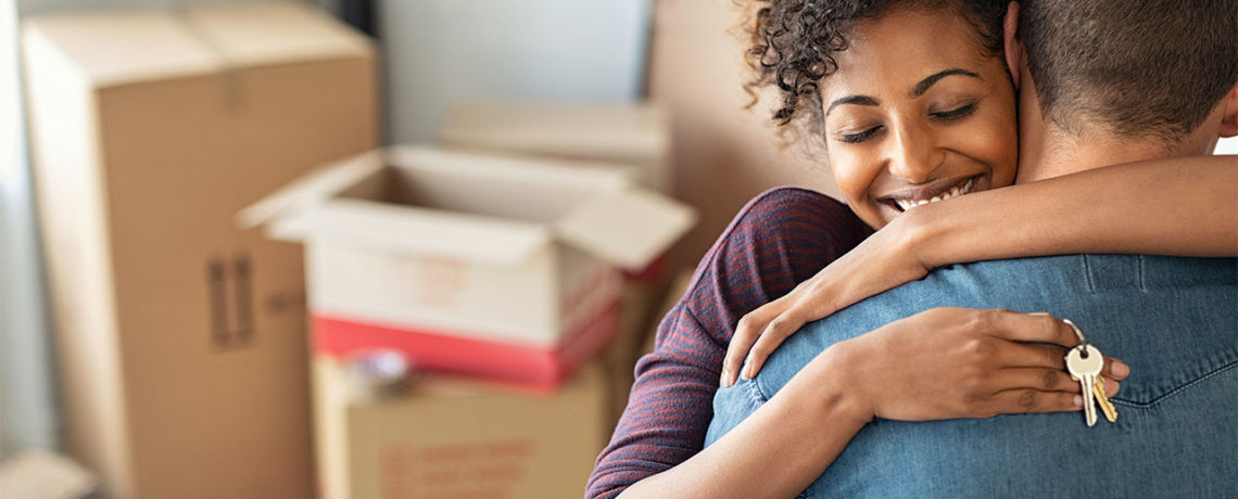 A man hugs a woman as she holds keys to their new home over his back in front of a stack of moving boxes.