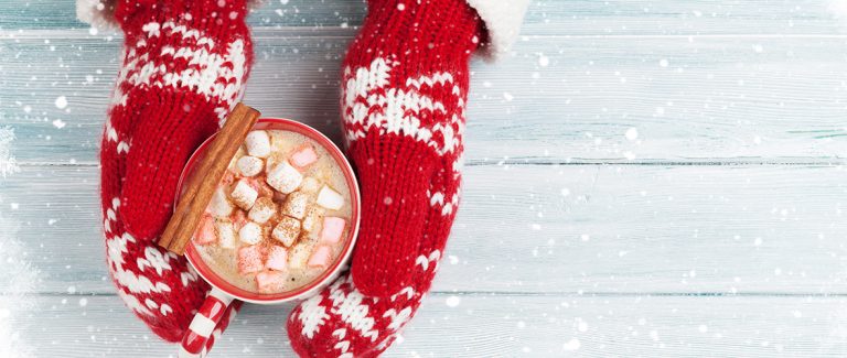 gloves on hands around a mug of hot chocolate with marshmallows and cinnamon sticks