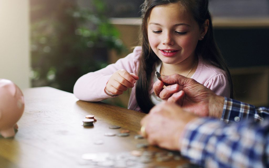 An older man teaches a young girl about financial literacy at a table with coins and a piggy bank