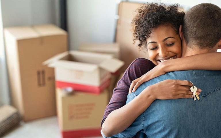 A man hugs a woman as she holds keys to their new home over his back in front of a stack of moving boxes.