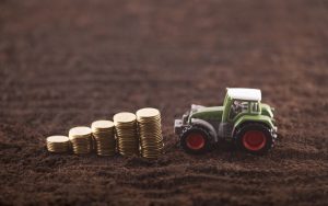 A miniature tractor and an ascending stack of coins rest on a plowed field.