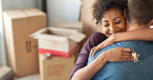 A man hugs a woman as she holds keys to their new home over his back in front of a stack of moving boxes.